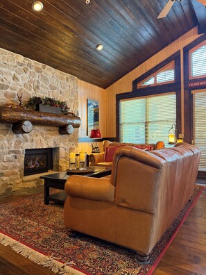 A cozy living room with a stone fireplace, leather sofa, and a large window with blinds. - Cozy Ski Chalet,  Mountain Views and short walk to base of Deer Valley! (Park City)