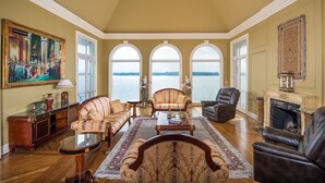 A spacious living room with a view of a lake, featuring a fireplace, leather sofas, and a large painting. - The Retreat with  Wilson Dam view & Water Aces (Florence)