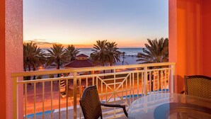 Un balcón con una mesa y sillas de vidrio, con vista a una playa con palmeras y un atardecer.