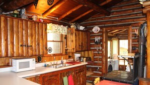 A rustic cabin kitchen with wooden walls, a central island, a microwave, a sink, and a wood stove. - Storybook Cottage in the Heart of the Big Horn Mountains (Story)