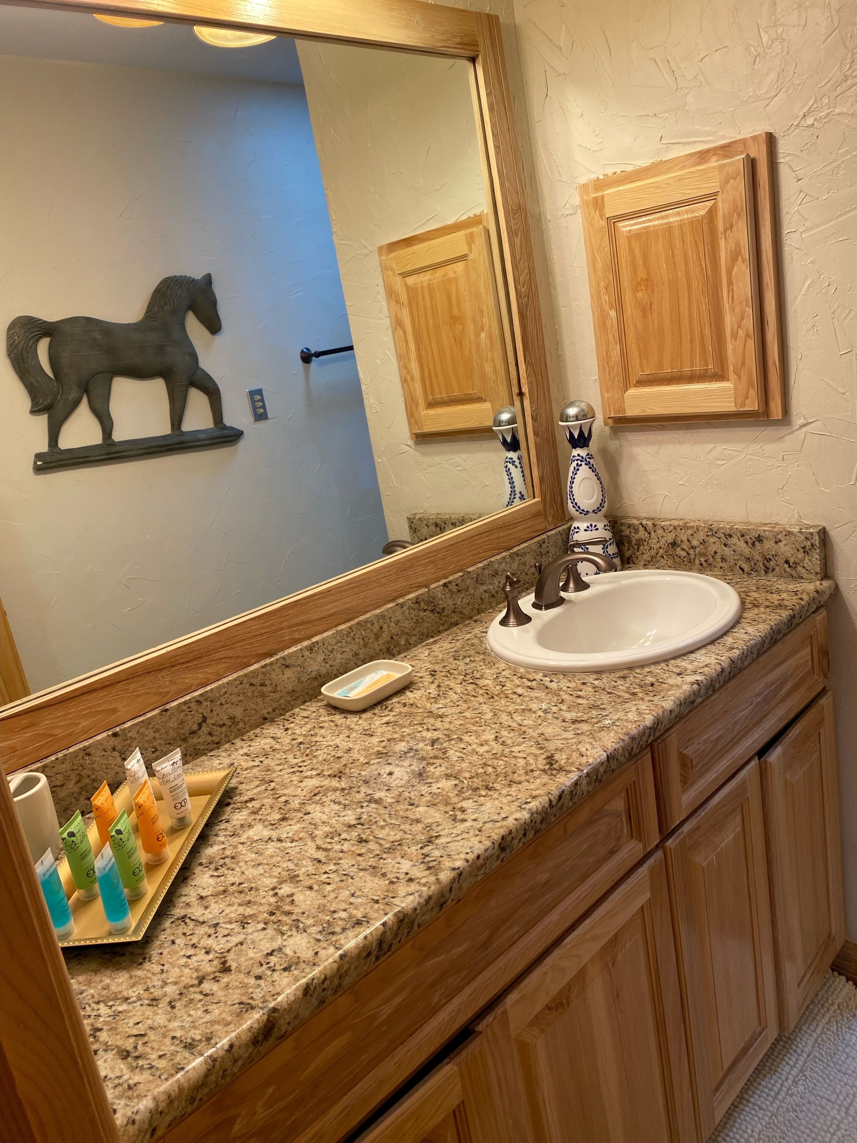 A bathroom with a granite countertop, a white sink, wooden cabinets, a mirror, and toiletries on a tray.