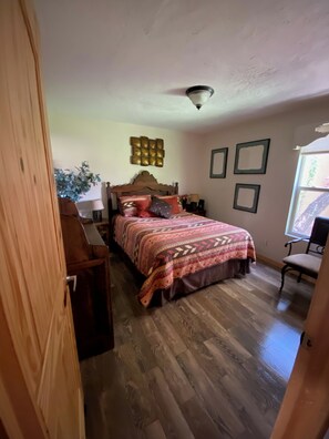 A bedroom with a wooden bed, a ceiling fan, a wall-mounted light fixture, and a window with a view of greenery. - Sandia Heights Getaway_VIEWS, UPSCALE, 3BR (Albuquerque)