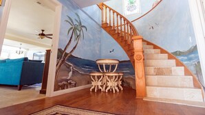Una sala de estar con escalera de madera, un mural con escena de playa y un sofá azul.