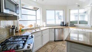 Cocina moderna con estufa a gas, electrodomésticos de acero inoxidable, armarios blancos y amplias ventanas con vistas a la playa.