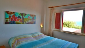 A bedroom with a bed, a window with curtains, a wall-mounted lamp, and a colorful painting. - Villa Porticciolo (Rio)