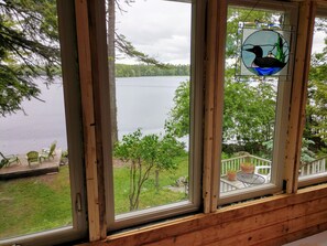 Una cabaña de madera con un ventanal grande que da a un lago, una terraza con sillas y una puerta de cristal decorada con un pájaro.