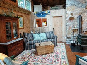 Interior acogedor de una cabaña con sofá de madera, un reposapiés de mimbre, un tapete estampado de flores, una pared de piedra y un letrero que dice 'Evening Cottage'.