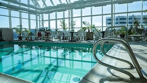 Piscina interior com paredes de vidro transparente, rodeada por espreguiçadeiras e plantas em vasos.