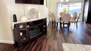 A modern living room with a dark wood floor, a flat-screen TV mounted on the wall, a dark wooden entertainment center, and a dining area with patterned chairs and a chandelier. - Beautifully, Renovated Home in Quiet Cul de Sac! Close to EVERYTHING!  (Oldsmar)