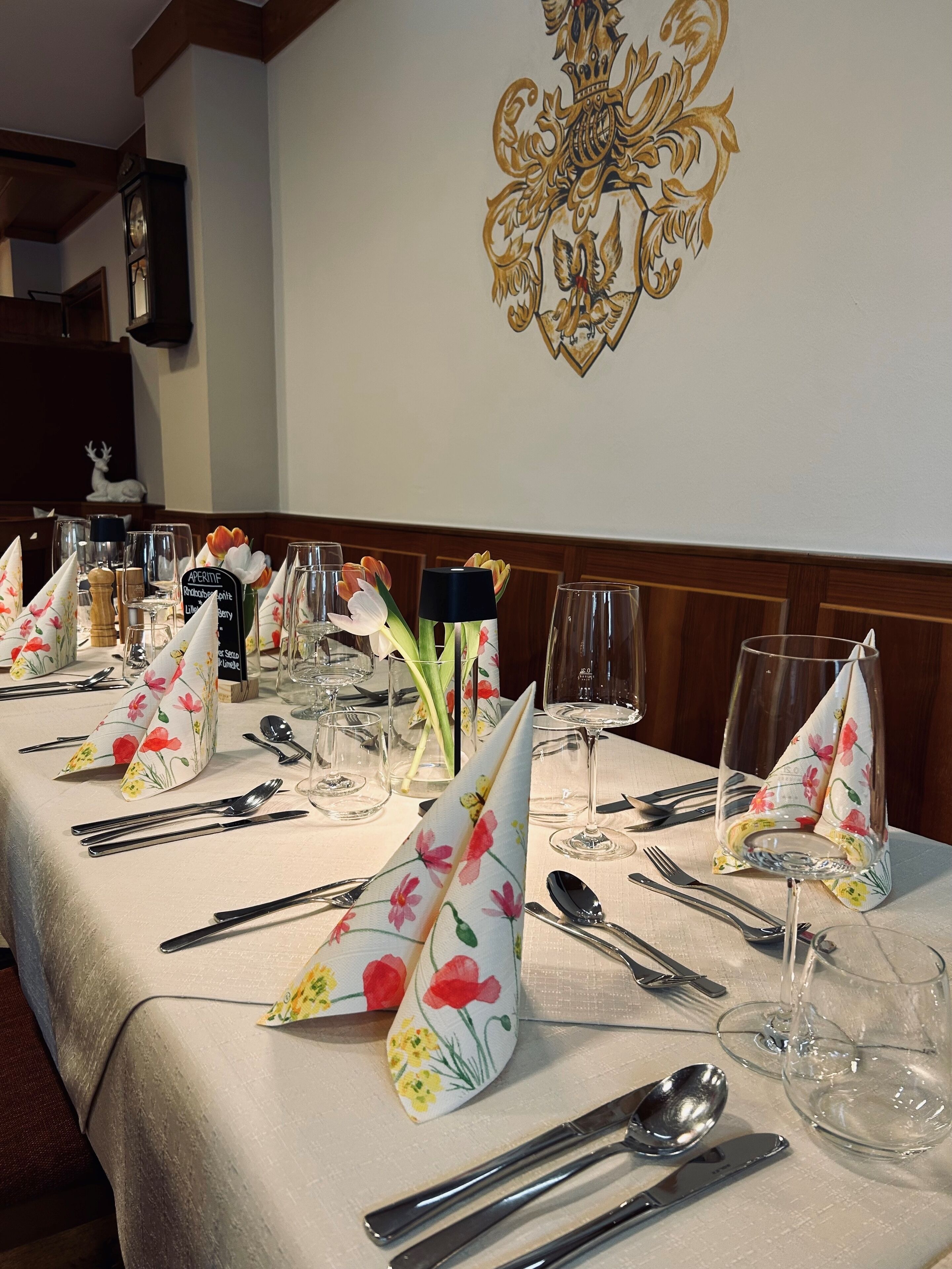 A dining table set with floral napkins, silverware, and wine glasses, with a decorative emblem on the wall behind.