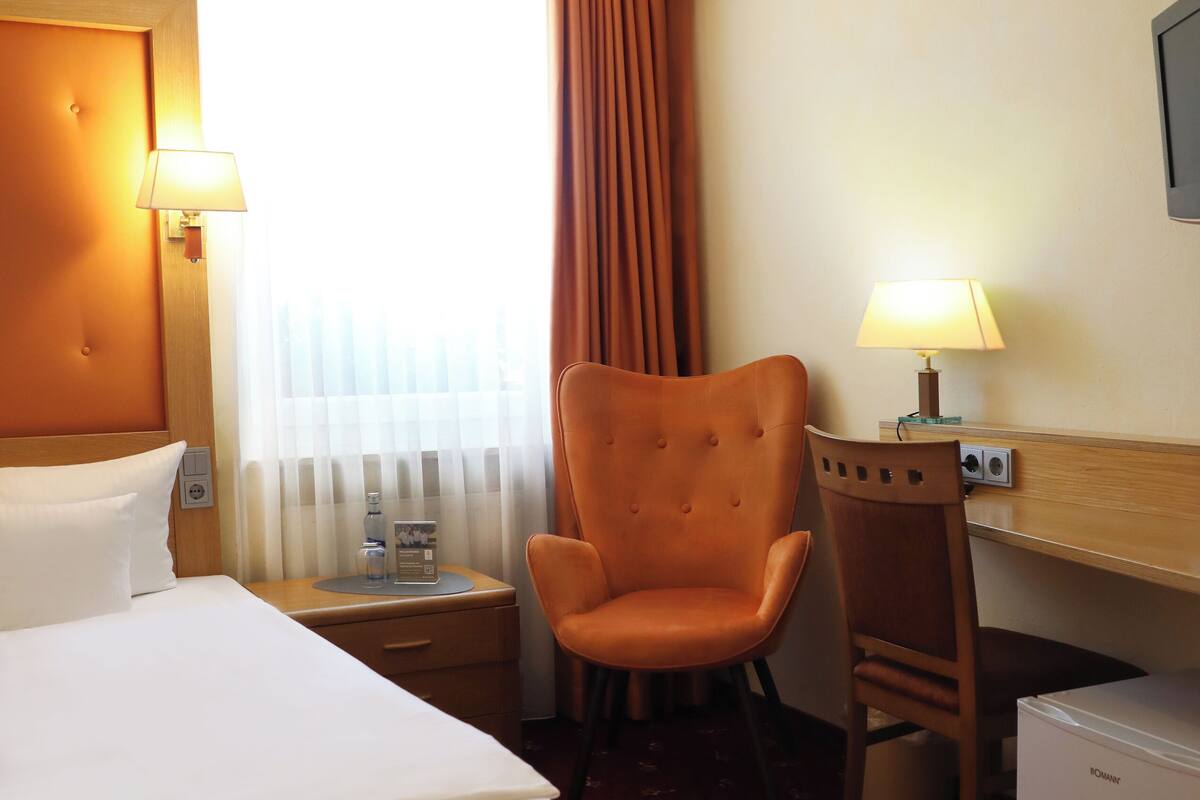A hotel room with a large bed, a chair, a desk with a lamp, and a small refrigerator.