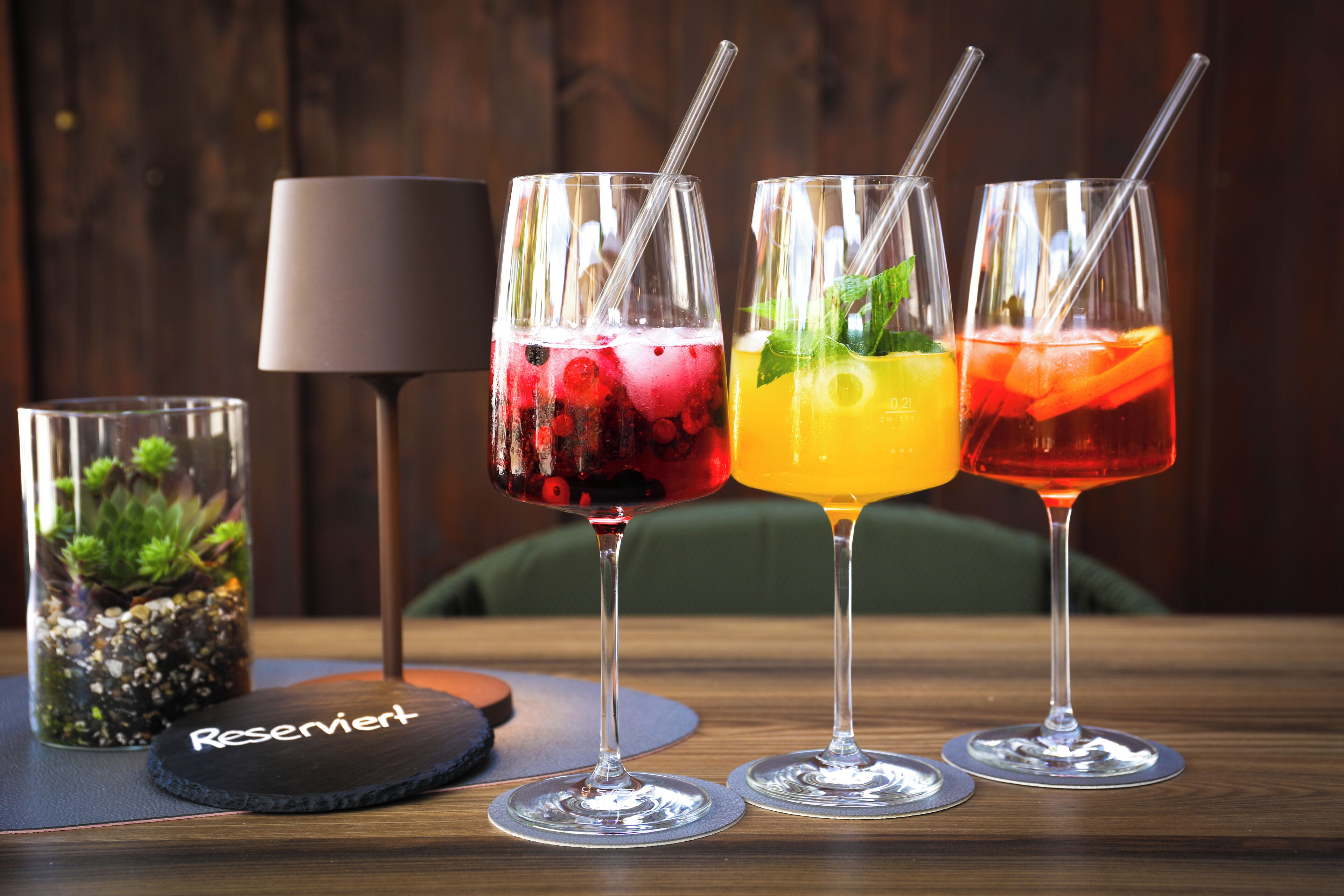 Three glasses of beverages with straws, a small glass with a plant, and a lamp on a table.