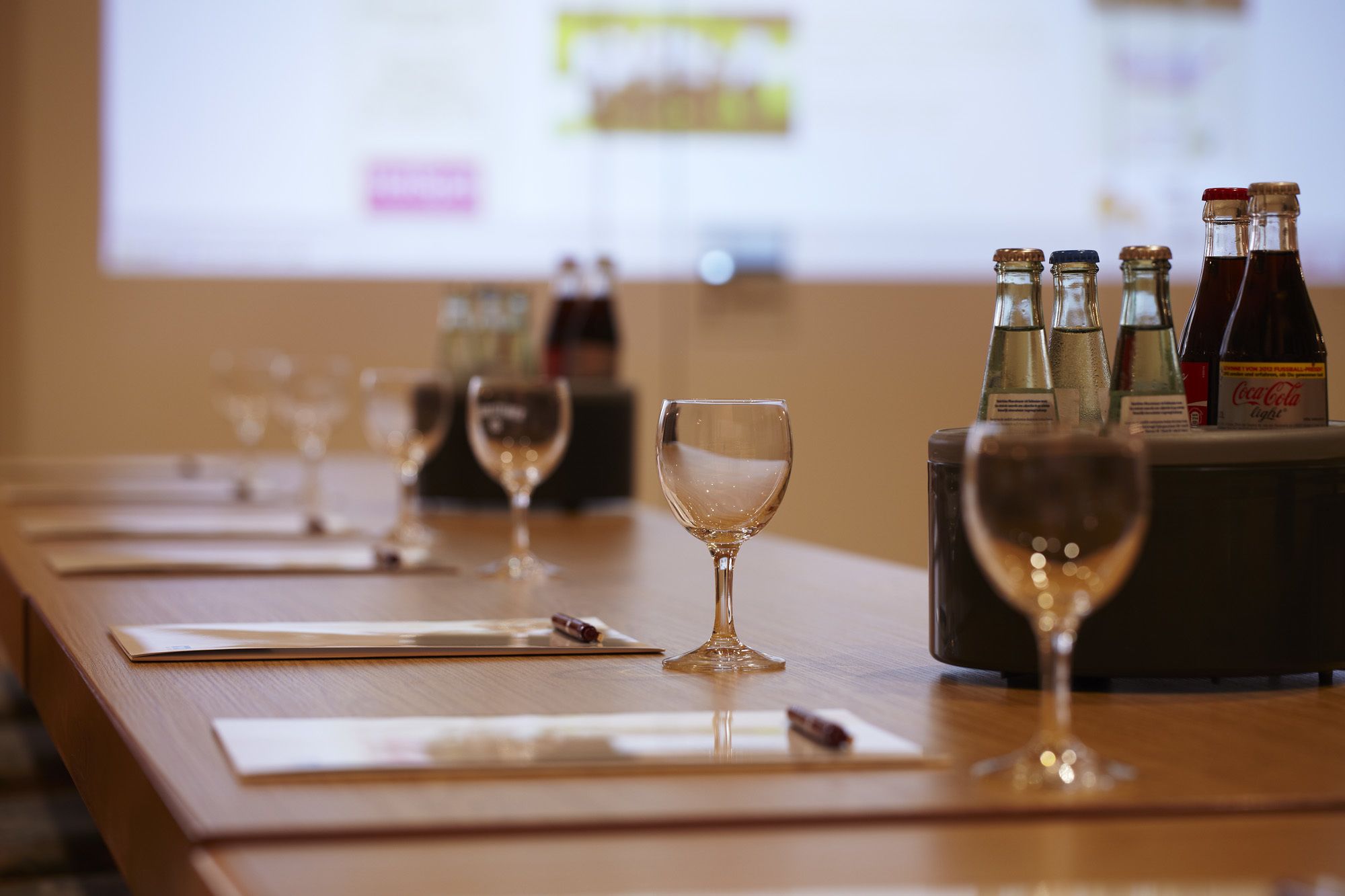Una mesa con copas y botellas de vino, una pantalla de proyector al fondo y superficie de madera.