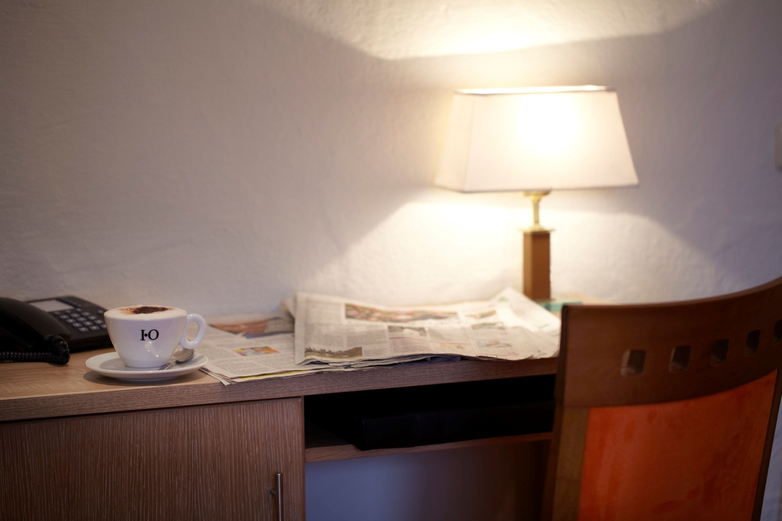 A hotel room with a desk, a cup of coffee, a lamp, and a chair.