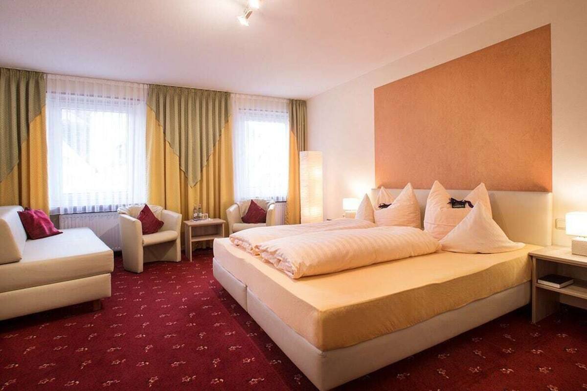 A hotel room with a large bed, two armchairs, a nightstand, a lamp, and a large window with curtains.
