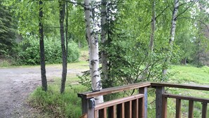 Un patio in legno con ringhiera in metallo, circondato da alberi e un sentiero sterrato.