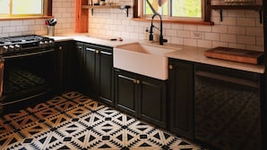 A kitchen with wooden beams, a patterned floor, a stove, and a window with a view. - The Frontenac: A Lakeside Cabin Retreat with Sauna (Godfrey)