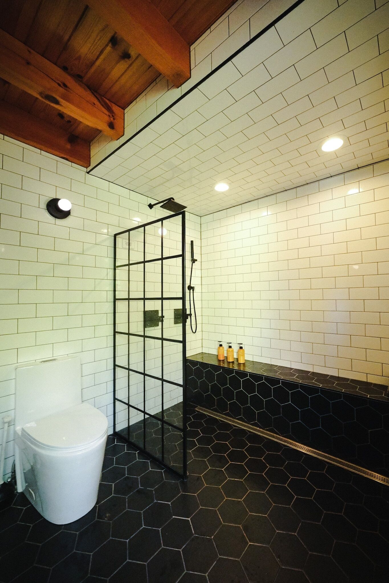 A bathroom with a white tiled wall, a black hexagonal floor, a wooden ceiling, a glass shower enclosure, and a white toilet.