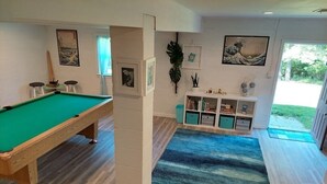 Una mesa de billar en una habitación con alfombra azul, suelo de madera y una estantería con diversos objetos.