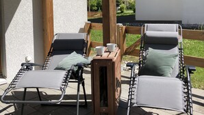A wooden pergola with two lounge chairs and a table, a small wooden table with cups, and a view of a mountain and a house in the background.