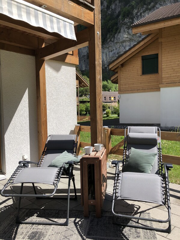 Une pergola en bois avec deux fauteuils et une table, une petite table en bois avec des tasses, et une vue sur une montagne et une maison au loin.