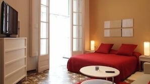 A hotel room with a bed, a television on a cabinet, a small round table, and a red rug. - 3BR/3BA Painted in White Apartment with Balcony. Free luggage storage service. (Barcelona)