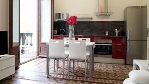 A modern kitchen with a dining area, featuring a tiled floor, a white dining table with chairs, a vase with red flowers, and a refrigerator. - 3BR/3BA Painted in White Apartment with Balcony. Free luggage storage service. (Barcelona)
