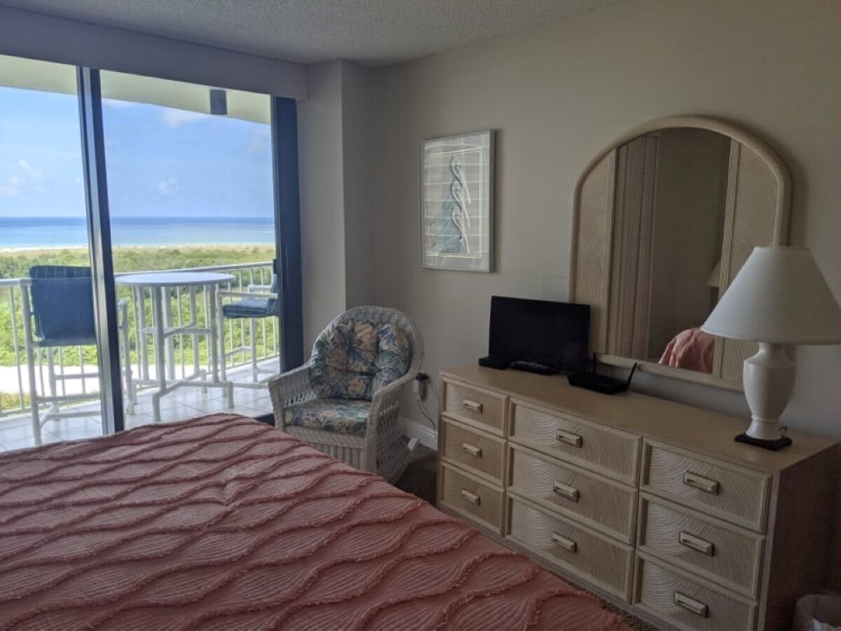 A bedroom with a bed, a dresser, a chair, a television, and a balcony with a view of the ocean.