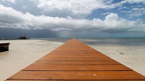 A wooden pier leading to a beach with a thatched-roof structure in the distance.