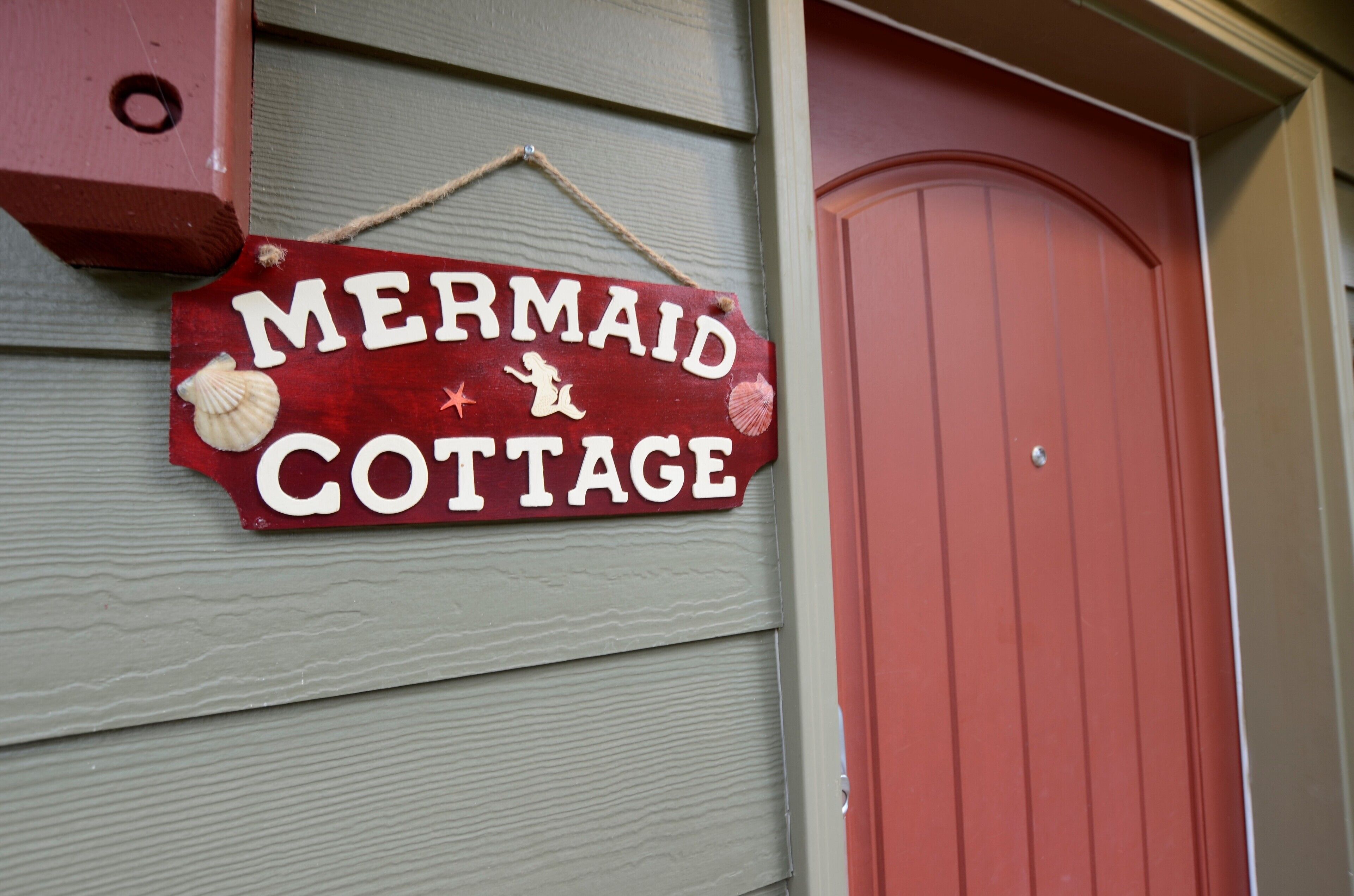Een houten bord met de tekst 'Mermaid Cottage' en decoratieve schelpen.
