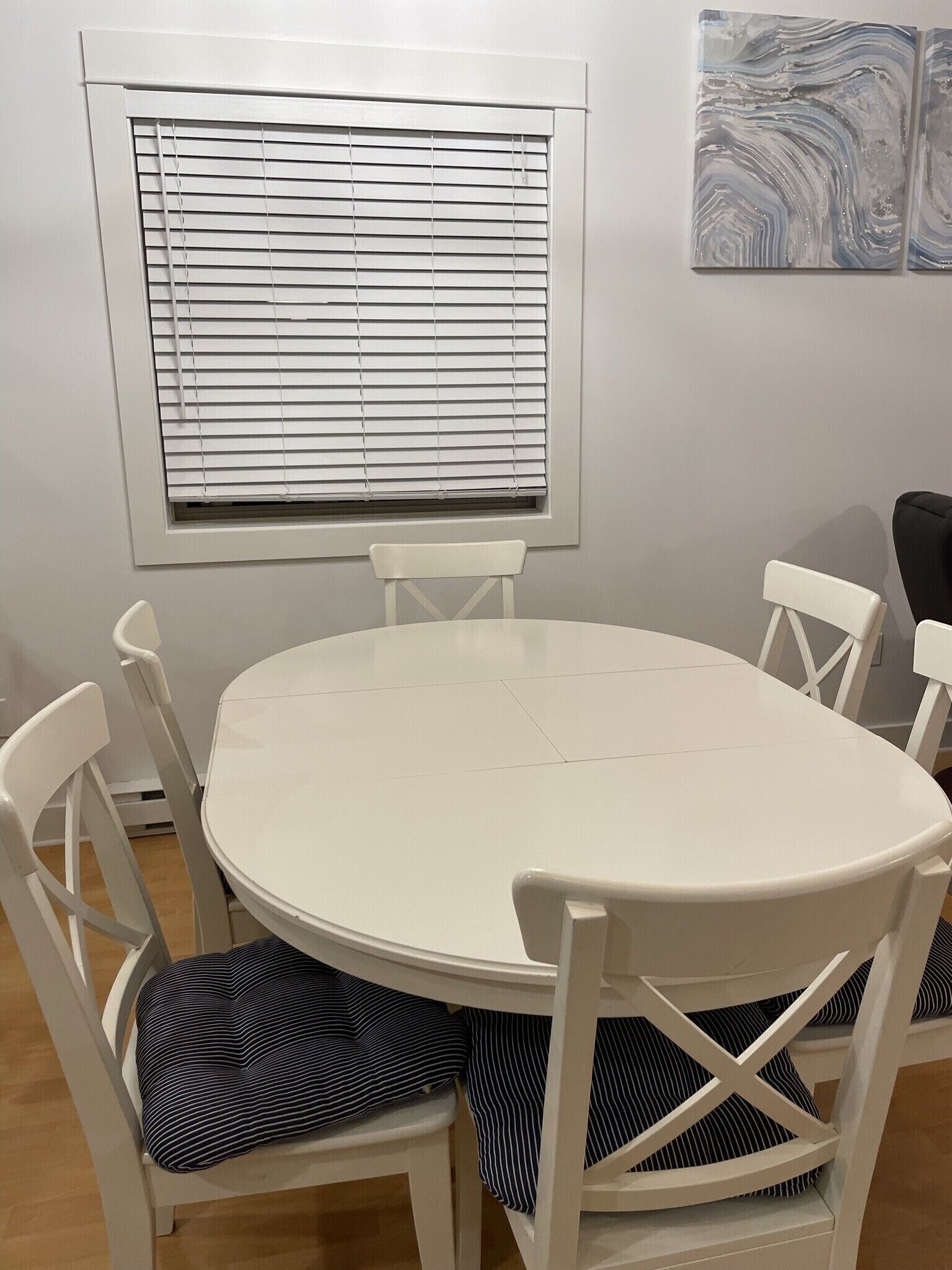 Una mesa redonda blanca con cuatro sillas blancas, cada una con un cojín a rayas. Encima de la mesa, hay una ventana con persianas.