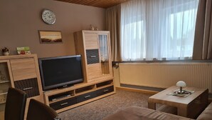 A living room with a television, wooden furniture, a sofa, a table, and a window with curtains. - Verwöhn-Pension Wiesengrund (Bischofsgruen)