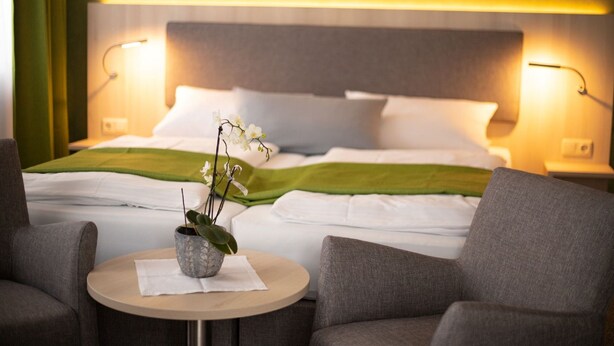 A hotel room with a neatly made bed, two bedside lamps, a small round table with a plant, and two armchairs. - Verwöhn-Pension Wiesengrund (Bischofsgruen)