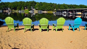 A lakeside view with green and blue Adirondack chairs on a sandy beach, boats docked in the background, and a forested shoreline. -  Remodeled Ledges Lakefront 2BR Condo! No Steps. Walk-In Unit w/ WiFi. (Osage Beach)