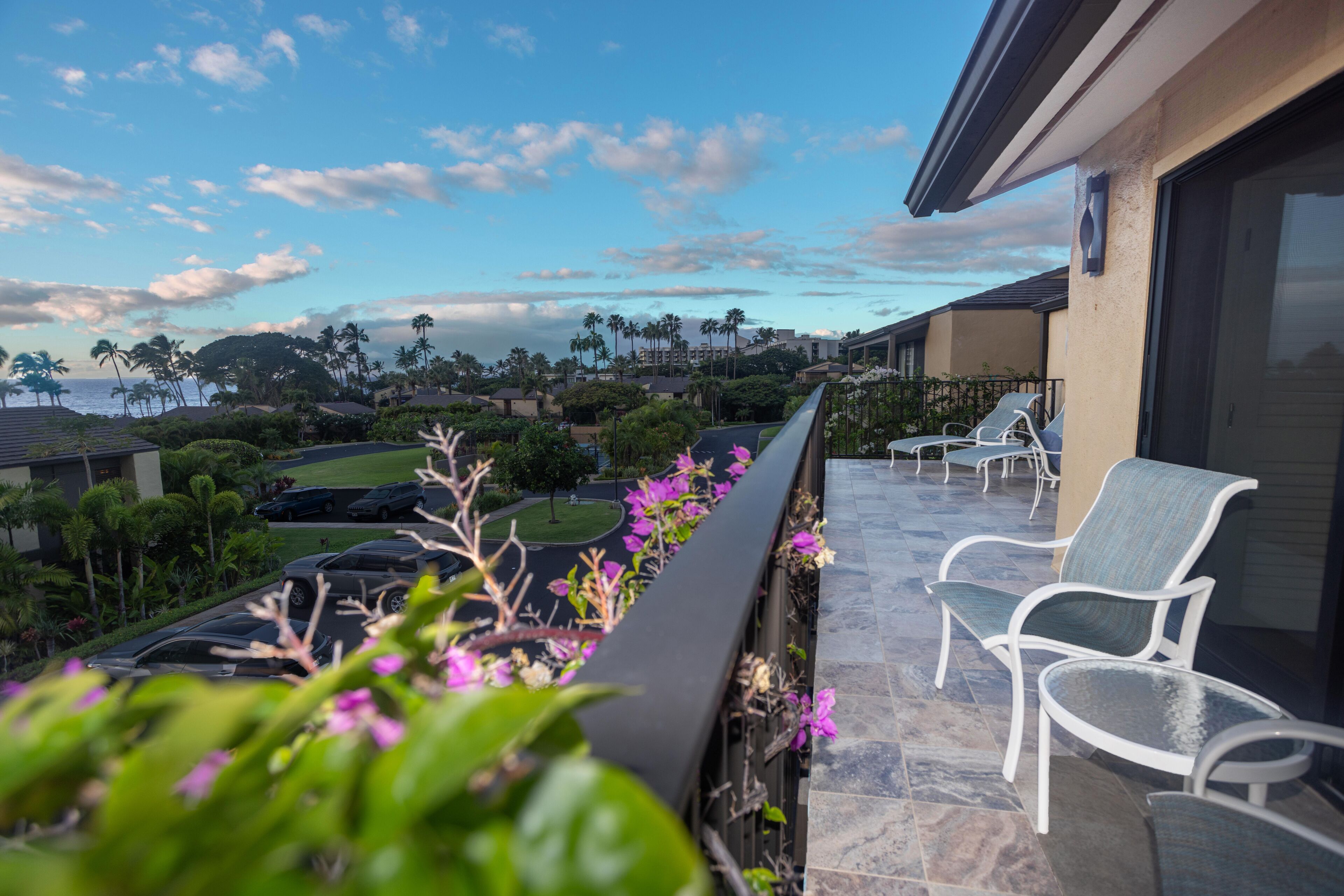 A balcony with white chairs, a view of the ocean, and palm trees.