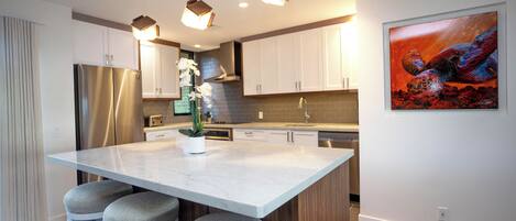 A modern kitchen with a marble island, bar stools, stainless steel appliances, and a large painting on the wall.