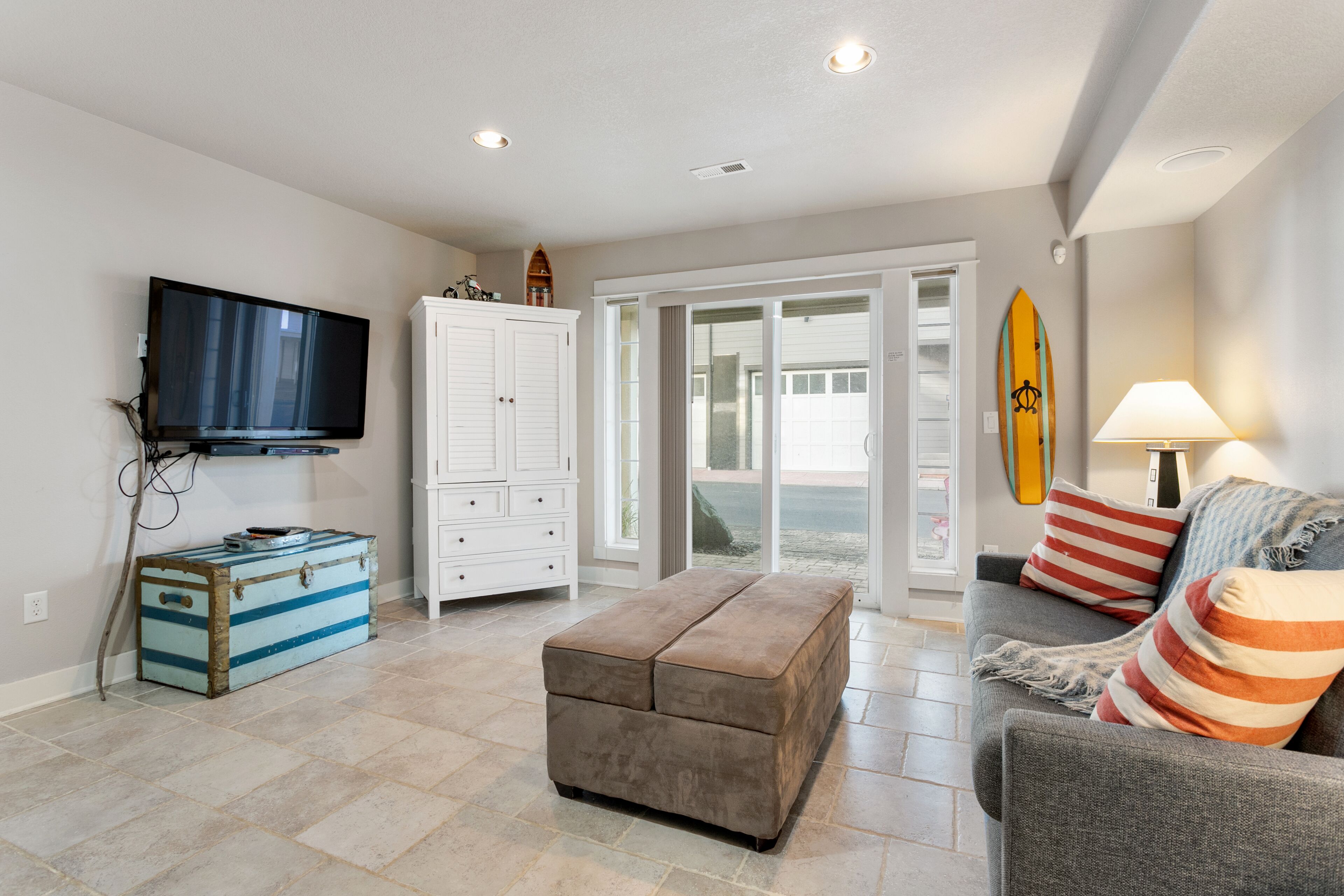 A living room with a flat-screen TV, a storage chest, a cushioned bench, a sofa with striped pillows, and a surfboard on the wall.