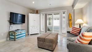 A living room with a flat-screen TV, a storage chest, a cushioned bench, a sofa with striped pillows, and a surfboard on the wall.