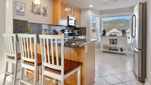 A modern kitchen with wooden cabinets, a central island with bar stools, a stainless steel refrigerator, and a large window offering a view of the outdoors.