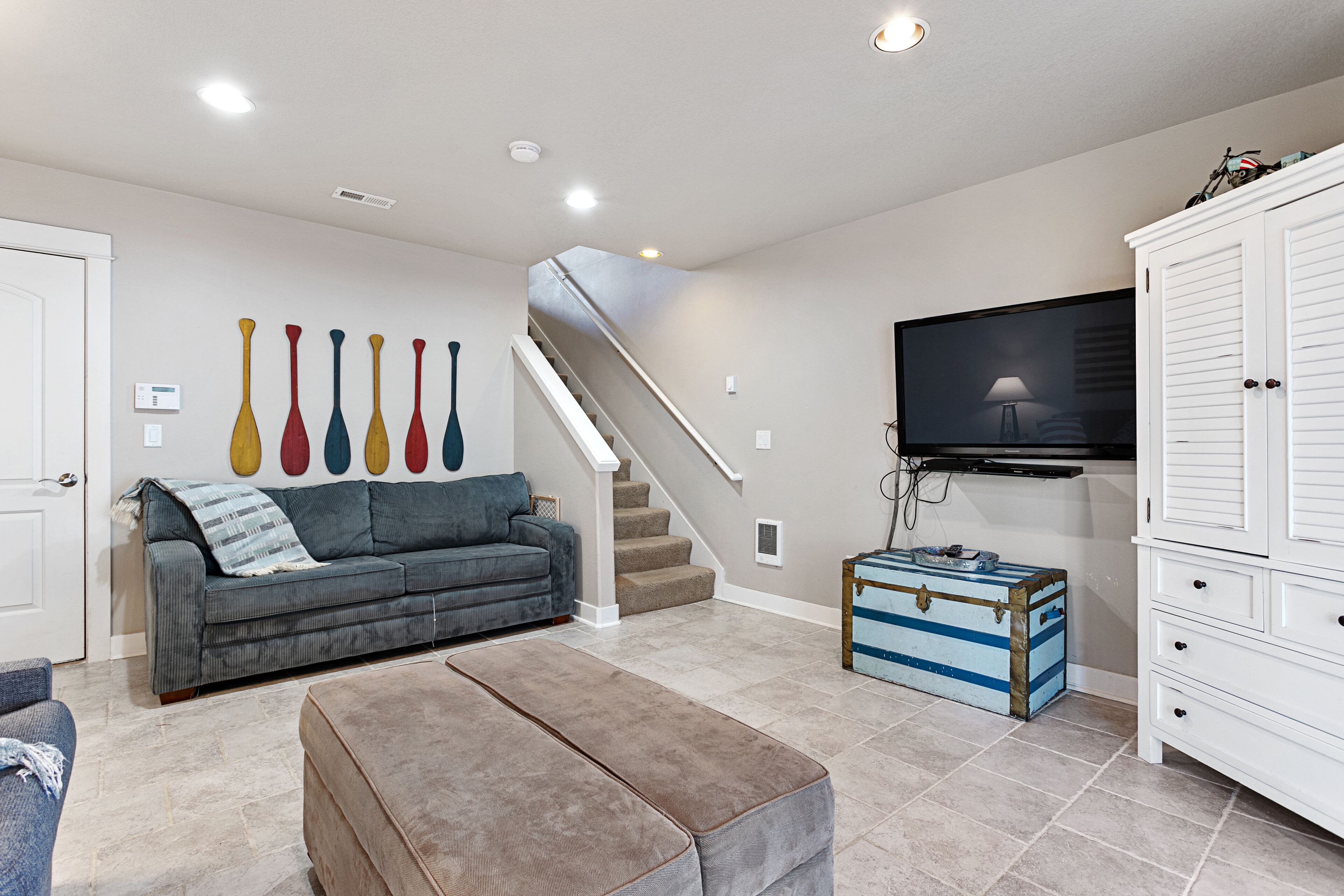 A living room with a grey sofa, a brown ottoman, a staircase, a television, and a white cabinet.