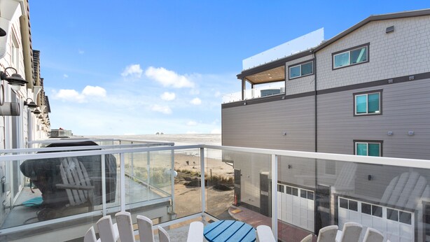 A balcony with white Adirondack chairs and a small blue table, overlooking a beach and adjacent buildings.