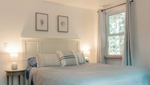 A bedroom with a bed, two framed pictures on the wall, a bedside table with a lamp, and a window with curtains. - Charming Cape Cod cottage on Depot Pond & bike trail with pond access (Eastham)