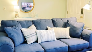 A living room with a blue sofa, white coffee table, and a television on the wall. - Charming Cape Cod cottage on Depot Pond & bike trail with pond access (Eastham)