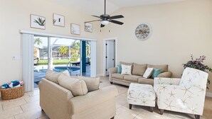 Ein Wohnzimmer mit einem beigefarbenen Sofa, einem passenden Sessel und einem Couchtisch. Es gibt einen Deckenventilator, eine Wanduhr und ein großes Fenster mit Blick auf Palmen.