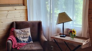A cozy cabin room with a wooden chair, a small table with a lamp and a vase of sunflowers, and a window with sheer curtains. - "Elk's Run"  Rustic Cabin Hidden Among The Pines At Falls Creek. (Condon)