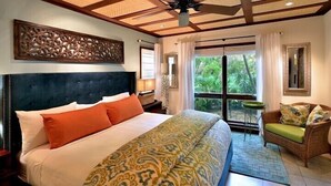 Ein Schlafzimmer mit einem großen Bett, einem Deckenventilator, einer dekorativen Wandverkleidung, einem Fenster mit Vorhängen und Blick ins Grüne.