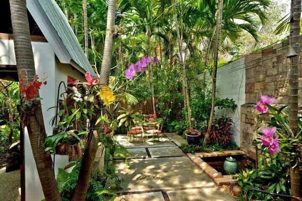 Relax in a serene garden setting surrounded by lush plants and flowers, including orchids, with a seating area for dining or relaxation.