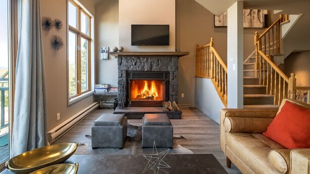 A living room with a stone fireplace, a flat-screen TV above it, a staircase with wooden railings, and a large window with curtains. - Tremblant Prestige - Clairiere 404 (Mont-Tremblant)