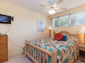 A bedroom with a bed, a wooden dresser, a ceiling fan, a window with blinds, and a television mounted on the wall.