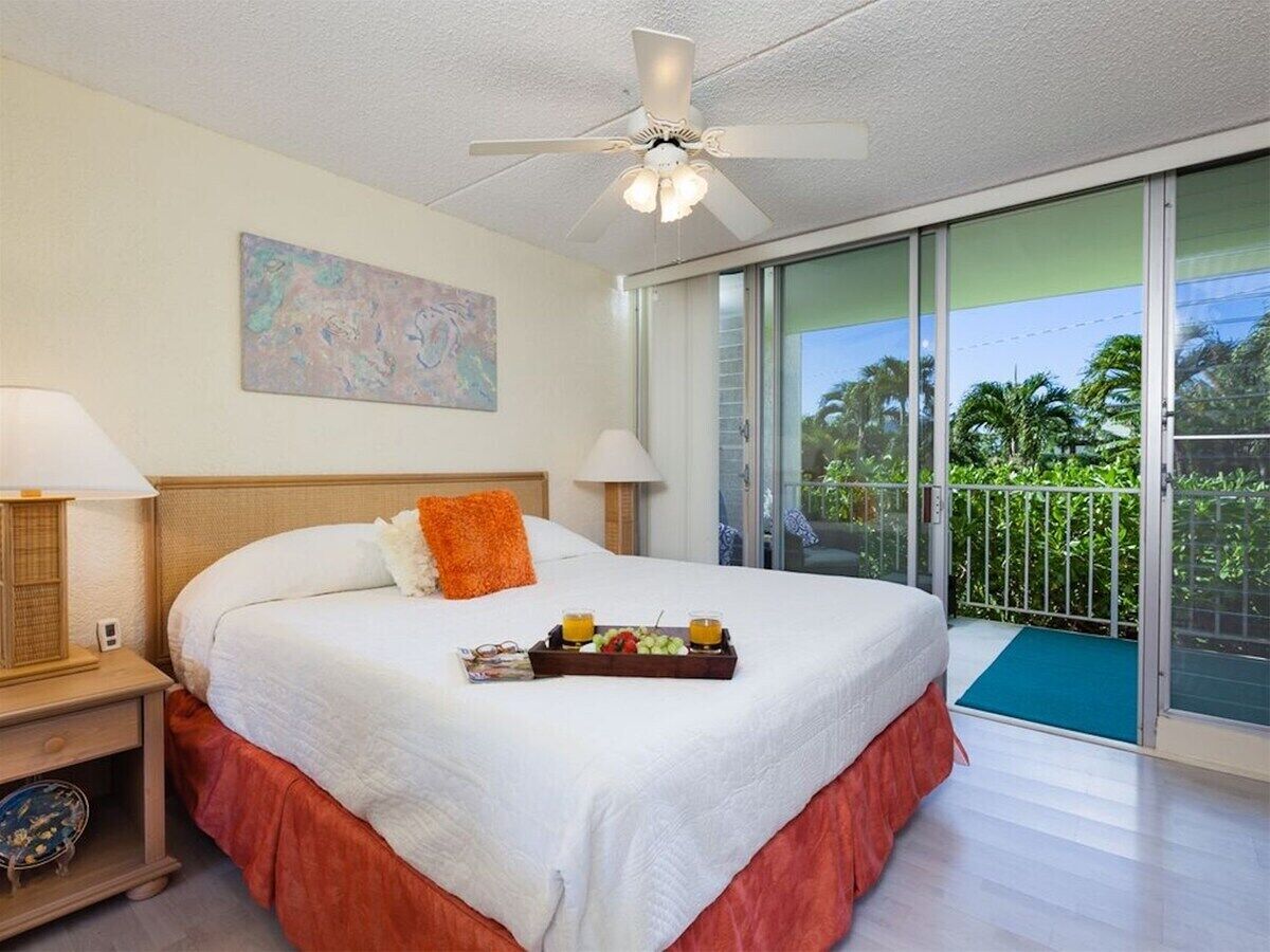A hotel room with a bed, a nightstand, a ceiling fan, a window with a view of greenery, and a balcony with a railing.